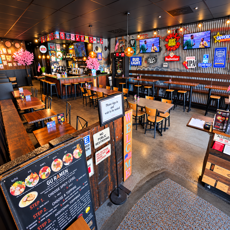 Colorful Japanese ramen restaurant interior with communal tables, bar seating, neon beer signs, televisions, and cherry blossom decorations.
